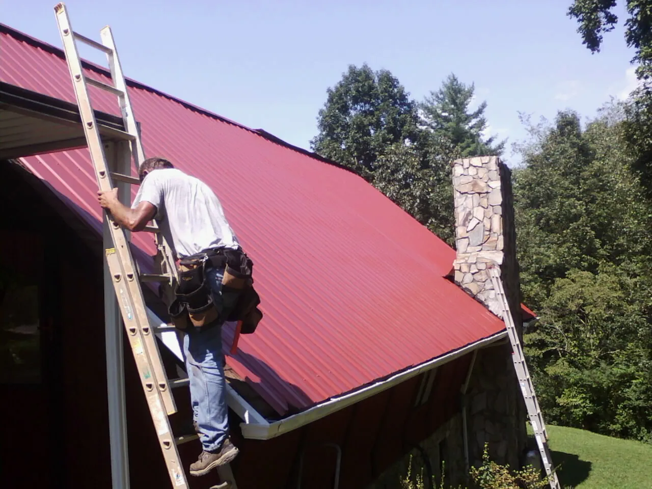 Roofer on a ladder beside a red metal roof and stone chimney — Soffit & Fascia Repair by Roofing Brothers in Fort Belvoir
